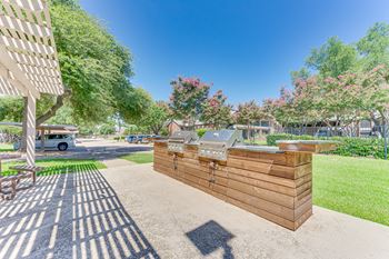 A wooden structure with a grill on top is situated in a sunny outdoor area with trees and a car in the background.
